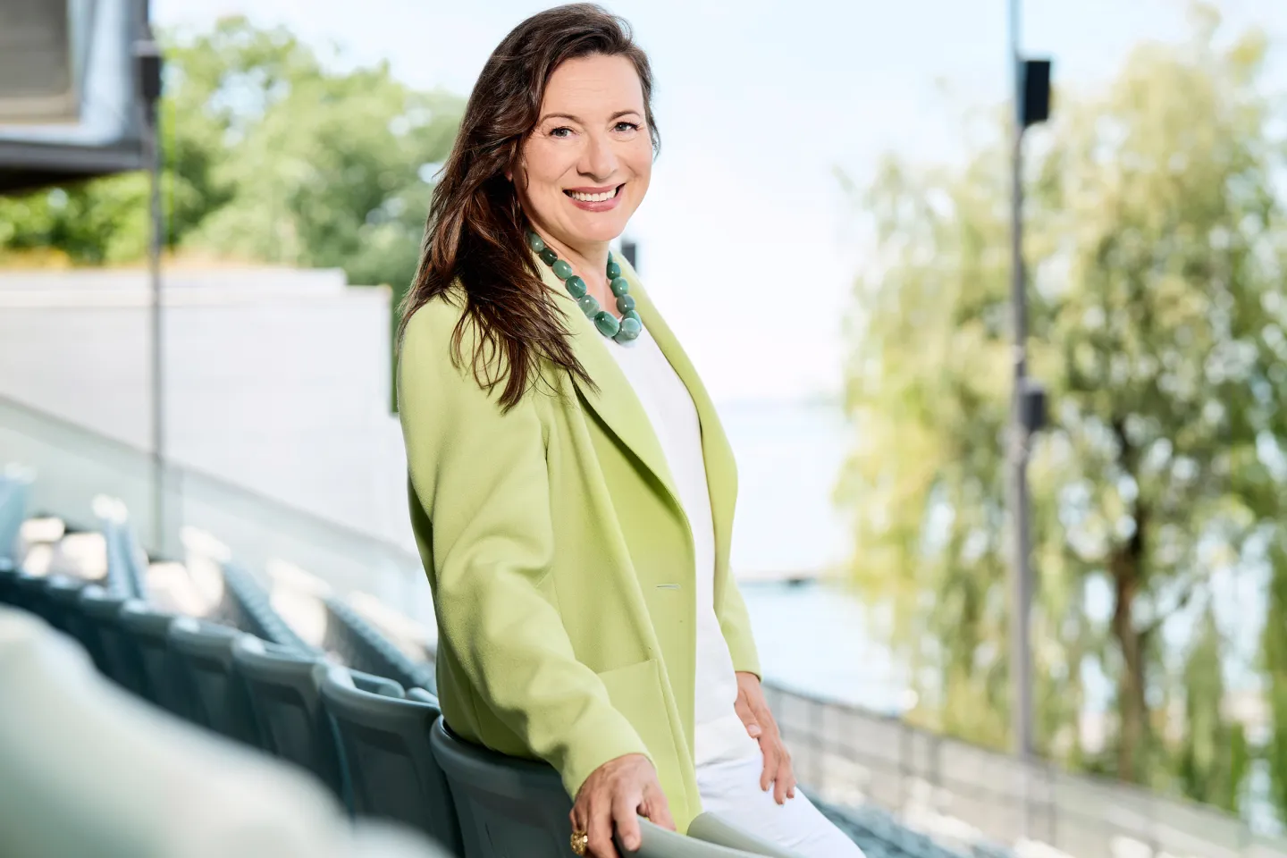 © Bregenzer Festspiele / Anja Köhler Lilli Paasikivi, Intendantin der Bregenzer Festspiele, lächelnd im Freien mit grüner Jacke und weißem Outfit vor verschwommenem Hintergrund mit Bäumen und Wasser