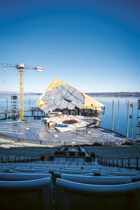 © Bregenzer Festspiele / Kristina Becker An open-air stage with rows of seats in front of a stage featuring a large, circular set piece and a crane by the lakeside under a clear sky.