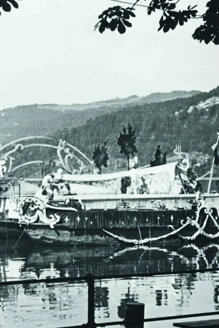 © Stadtarchiv Bregenz / Anni Sonnleitner Black-and-white photo of a boat decorated with ornaments on a lake, with forested hills in the background and a tree in the foreground.