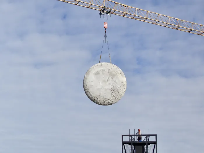 © Bregenzer Festspiele / Pia Kiker Runder, kraterübersäter Mond wird von einem Baukran an zwei Seilen über einem Gerüst mit einer Person gehoben.