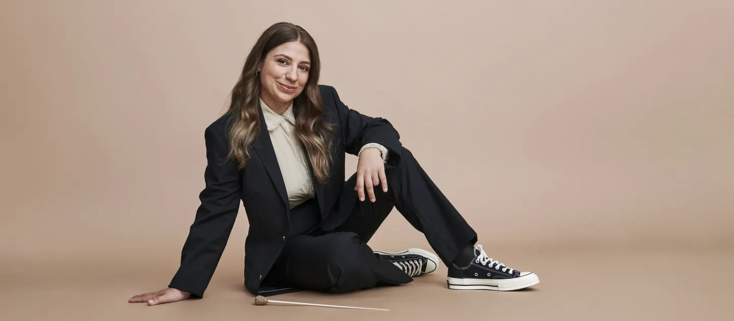 © Veikko Ka╠êhko╠ênen Frau sitzt auf dem Boden, trägt schwarzen Anzug, weiße Bluse und schwarze Sneakers, hält Dirigentenstab