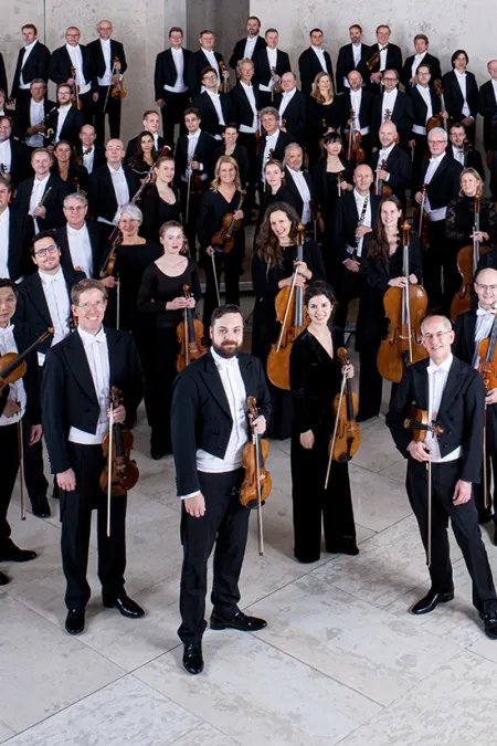 © Bregenzer Festspiele / Peter Rigaud Mitglieder der Wiener Symphoniker in formeller Konzertkleidung mit Instrumenten, auf einer Treppe in einem modernen Saal gruppiert