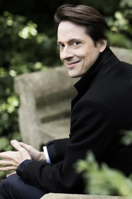 © Benjamin Ealovega Dirigent Leo McFall sitzt auf einer Steinbank im Grünen, trägt einen schwarzen Mantel und lächelt leicht zur Seite