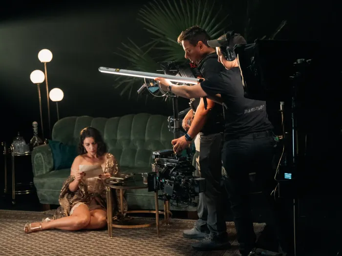 © Bregenzer Festspiele / Jonatan Langendorf Frau in goldfarbenem Kleid sitzt auf Teppich vor grünem Sofa, zwei Männer bedienen Filmkamera und Lichttechnik.